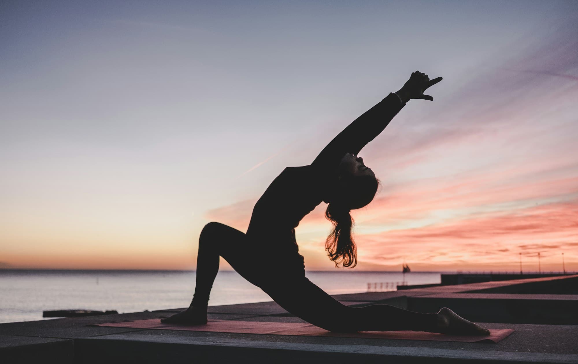 A person silhouetted in a low lunge stretch on a mat beside the sea at sunset
