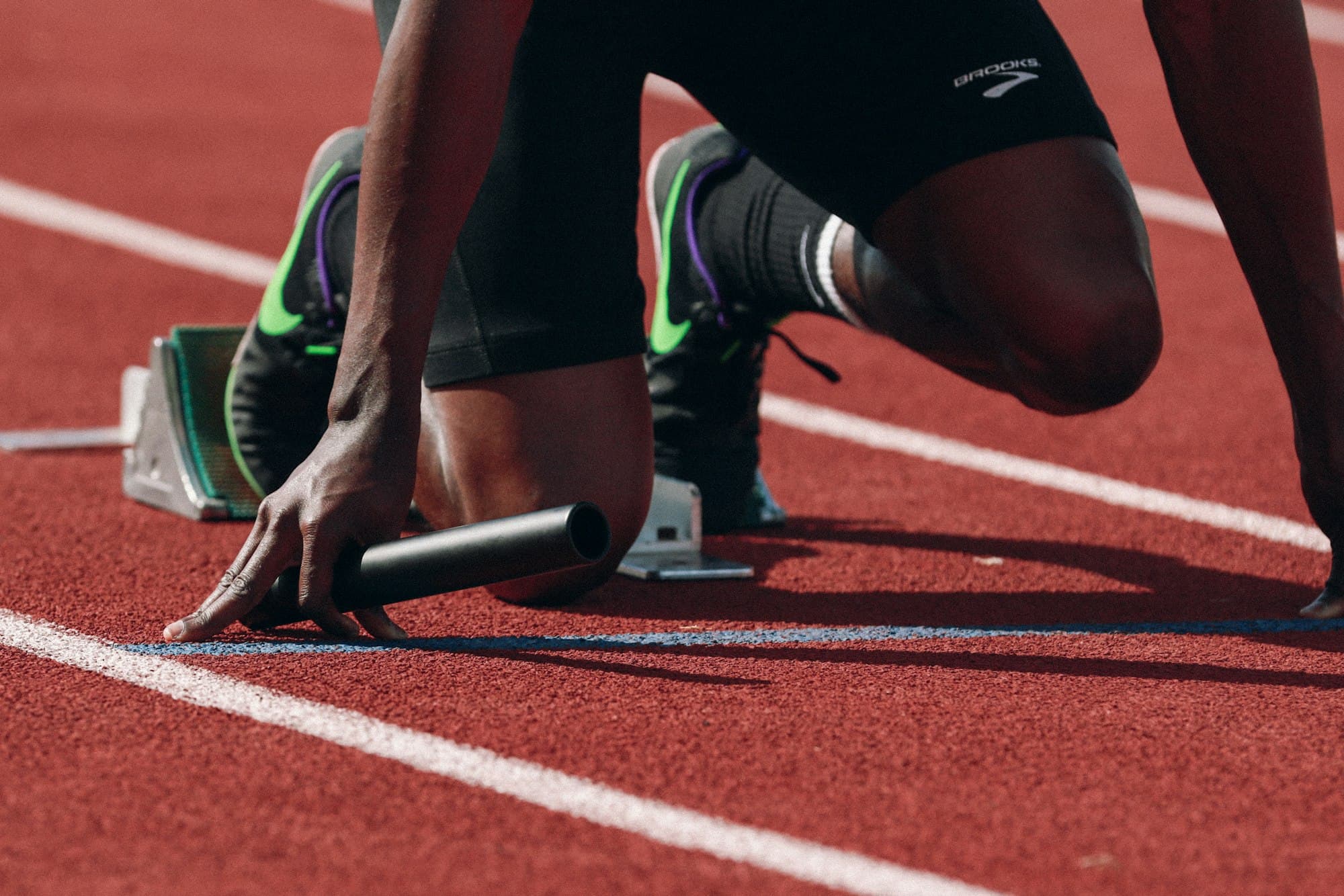 A sprinter set in the starting blocks on a red running track, holding a relay baton